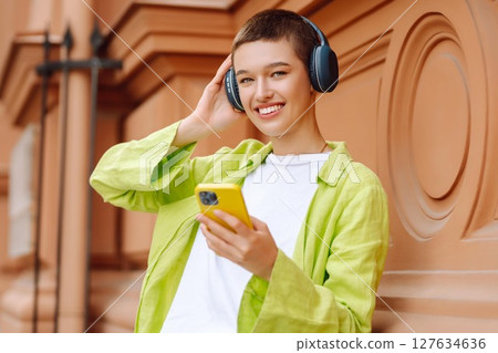 Woman wearing headphones posing outdoors, listens to music and walks along streets of a sunny city. Woman wearing headphones posing outdoors, listens to music and walks along streets of a sunny city. 127634636