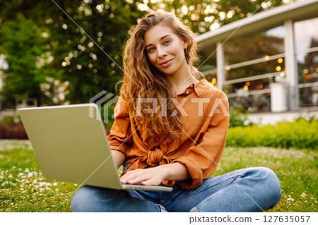 Portrait of a happy woman in sunny park on green lawn with a laptop. Freelance concept, technology. 127635057