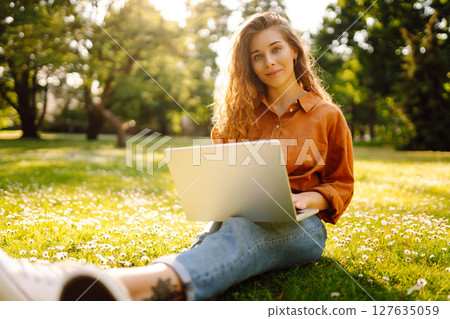 Portrait of a happy woman in sunny park on green lawn with a laptop. Freelance concept, technology. 127635059