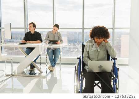 Accessible environment. African woman in chair for people with disability working with laptop in modern office. Diverse multiracial group of people colleagues working together. Young professional team 127635252