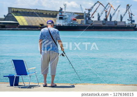 Senior man fishing near port waterfront, holding rod beside blue chair and bucket under sunlight. Concept of fishing, peaceful routine, retirement lifestyle by seaside 127636015