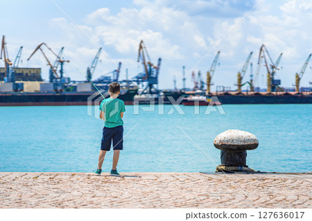 Boy fishing on sunny pier with industrial port in background, standing near turquoise water. Concept of fishing, childhood moments, peaceful outdoor leisure 127636017