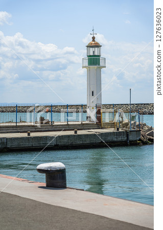 Old white lighthouse with green accents stands near dockside, fenced off at harbor under bright daylight. Concept of sea lighthouse, coastal safety, navigation structure 127636023