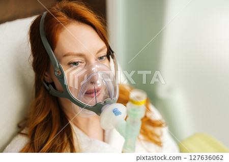 Close-up face female patient performing respiratory training, comfortably breathing with oxygen mask connected to respiratory device, promoting lung capacity and overall respiratory health, close-up. Close-up face female patient performing respiratory training, comfortably breathing with oxygen mask connected to respiratory device, promoting lung capacity and overall respiratory health, close-up. 127636062
