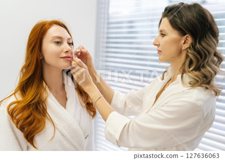 Female cosmetologist marking facial grid with white pencil, preparing redhead young woman client for cosmetic procedure in brightly lit aesthetic treatment room standing by window. Female cosmetologist marking facial grid with white pencil, preparing redhead young woman client for cosmetic procedure in brightly lit aesthetic treatment room standing by window. 127636063