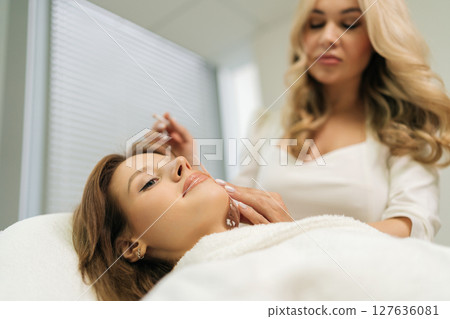 Closeup of skilled makeup artist marking facial contours with white pencil before cosmetic procedure, working in bright beauty salon environment near female client. Concept of beauty treatment. 127636081