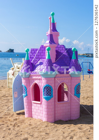 Pink plastic fairy-tale castle on beach sand. Seaside playground with loungers, umbrellas, and mountains in background. 127636542