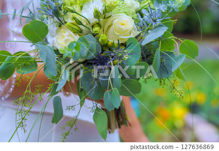 Bridal bouquet filled with white roses, greenery, purple accents held by bride in garden. 127636869