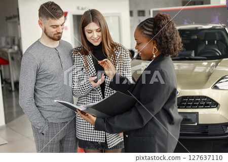 Young couple listening a sales agent about a car buying. Black female sales agent showing a documents and papers. Brunette caucasian man and woman is going to buy a car. Young couple listening a sales agent about a car buying. Black female sales agent showing a documents and papers. Brunette caucasian man and woman is going to buy a car. 127637110