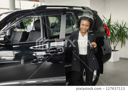 Young and cheerful woman enjoying new car while standing behinde. Black woman buy a new car. Girl wearing black costume. 127637113