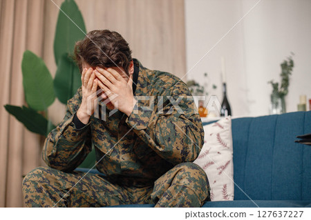 Portrait of young sad, desperate military man sitting on a coach during therapy session. Soldier lower head and hide his face. Man wearing camouflage. Portrait of young sad, desperate military man sitting on a coach during therapy session. Soldier lower head and hide his face. Man wearing camouflage. 127637227