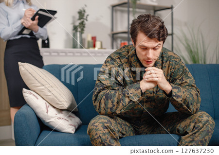 Man in military uniform sitting on a couch, woman psychiatrist standing behind. Soldier and psychiatrist talking during therapy session. Male warrior with ptds treating his mental disorder. Man in military uniform sitting on a couch, woman psychiatrist standing behind. Soldier and psychiatrist talking during therapy session. Male warrior with ptds treating his mental disorder. 127637230