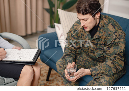 Cropped photo of a soldier and psychiatrist sitting on couch during therapy session. Man wearing military uniform. Male warrior with ptds talking to psychiatrist. 127637231