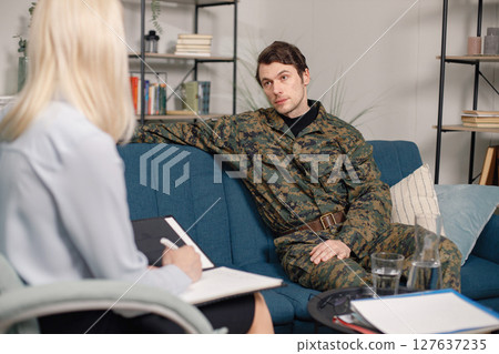 Soldier and psychiatrist sitting on couch during therapy session. Man wearing military uniform. Male warrior with ptds talking to psychiatrist. 127637235
