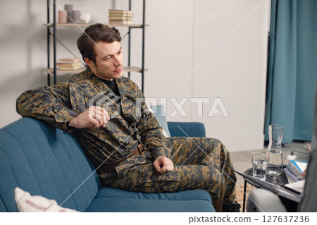 Cropped photo of a soldier sitting on couch during therapy session. Man wearing military uniform. Male warrior with ptds talking to psychiatrist. 127637236