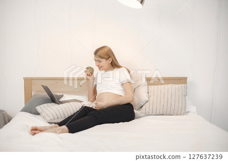Portrait of pregnant woman holding an apple while typing on a laptop. Pregnant woman lie on a bed in living room. Blonde woman wearing white t-shirt and black trousers. Portrait of pregnant woman holding an apple while typing on a laptop. Pregnant woman lie on a bed in living room. Blonde woman wearing white t-shirt and black trousers. 127637239