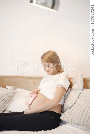 Portrait of pregnant woman holding a little baby rose socks. Pregnant woman lie on a bed in living room. Blonde woman wearing white t-shirt and black trousers. 127637241