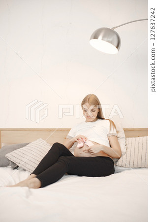 Portrait of pregnant woman holding a little baby rose socks. Pregnant woman lie on a bed in living room. Blonde woman wearing white t-shirt and black trousers. Portrait of pregnant woman holding a little baby rose socks. Pregnant woman lie on a bed in living room. Blonde woman wearing white t-shirt and black trousers. 127637242