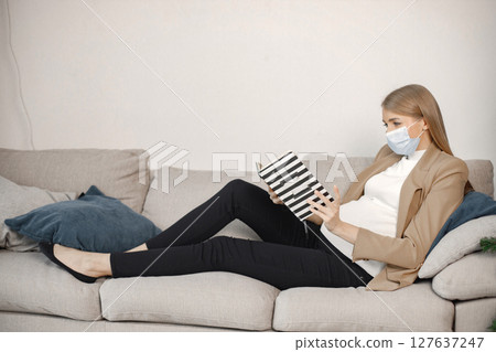 Portrait of pregnant woman holding a book and reading. Pregnant woman lie on a sofa in living room. Blonde woman wearing beige jacket and black trousers. 127637247