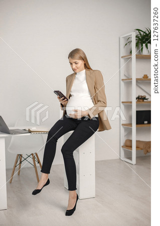 Portrait of pregnant woman using a phone and touching belly while sitting on desk in modern office. Pregnant businesswoman using a phone while sitting in her office. Blonde woman wearing beige jacket Portrait of pregnant woman using a phone and touching belly while sitting on desk in modern office. Pregnant businesswoman using a phone while sitting in her office. Blonde woman wearing beige jacket 127637260
