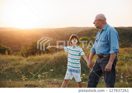 Photo of little boy and his grandfather walking in the field. Boy and grandfather spending time together at summer. Brunette boy wearing striped t-shirt and man blue shirt and glasses. Photo of little boy and his grandfather walking in the field. Boy and grandfather spending time together at summer. Brunette boy wearing striped t-shirt and man blue shirt and glasses. 127637261