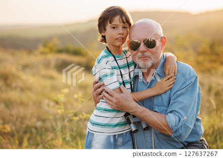 Photo of little boy and his grandfather standing and hugging in the field. Boy and grandfather spending time together at summer. Brunette boy and man wearing striped shirts. 127637267