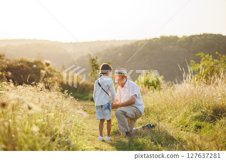 Backside photo of little boy and his grandfather standing and talking in the field. Boy and grandfather spending time together at summer. Brunette boy and man wearing striped shirts. Backside photo of little boy and his grandfather standing and talking in the field. Boy and grandfather spending time together at summer. Brunette boy and man wearing striped shirts. 127637281