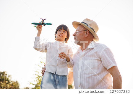 Portrait of little explorer and his grandfather playing with toy plane in a field. Boy and grandfather spending time together at summer. Brunette boy and man wearing striped shirts. 127637283
