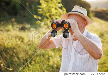 Portrait of a mature man with binoculars in a field. Bearded caucasian man looking through a binoculars in a field at summer. Man wearing striped t-shirt and a hat. 127637284