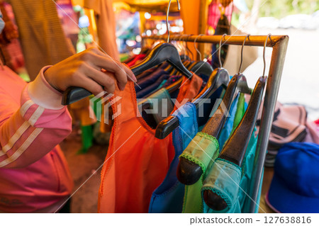Woman shopping for colorful traditional clothes at rural Thai street market where local vendors sell handmade garments to tourists during cultural travel experience in countryside Thailand Woman shopping for colorful traditional clothes at rural Thai street market where local vendors sell handmade garments to tourists during cultural travel experience in countryside Thailand 127638816