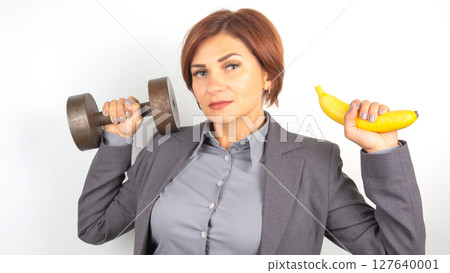 Happy beautiful business woman in a suit lifts a dumbbell and a banana in her hands Happy beautiful business woman in a suit lifts a dumbbell and a banana in her hands 127640001