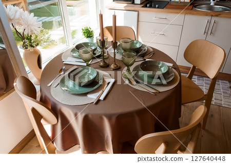 Cozy Dining Table Setting with Green Dishware, Candles, and Wooden Chairs in a Bright Minimalist Kitchen Interior Design, Earth Tones Cozy Dining Table Setting with Green Dishware, Candles, and Wooden Chairs in a Bright Minimalist Kitchen Interior Design, Earth Tones 127640448