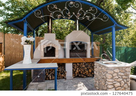 Outdoor Stone Pizza Oven with Decorative Wrought Iron Canopy and Blue Accents in a Lush Backyard Garden Setting. Outdoor Stone Pizza Oven with Decorative Wrought Iron Canopy and Blue Accents in a Lush Backyard Garden Setting. 127640475