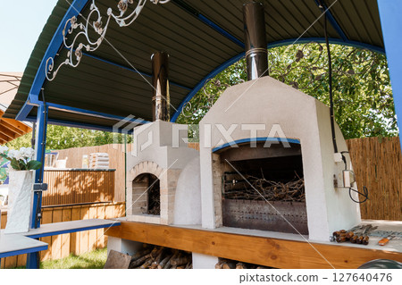 Outdoor Wood-Fired Pizza Oven with Smoker and Pergola Shelter: Rustic Backyard Cooking and Dining Setup in Summer Light Outdoor Wood-Fired Pizza Oven with Smoker and Pergola Shelter: Rustic Backyard Cooking and Dining Setup in Summer Light 127640476