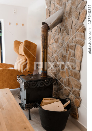 Cozy Interior with Stone Fireplace and Wood Stove. Rustic Living Room Featuring Orange Chair, Log Storage, Black Stove Pipe, and Warm Tones 127640835