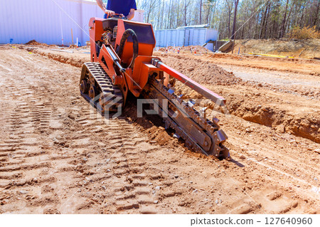 Heavy trenching machinery digs trench in soil for construction purposes on work site Heavy trenching machinery digs trench in soil for construction purposes on work site 127640960