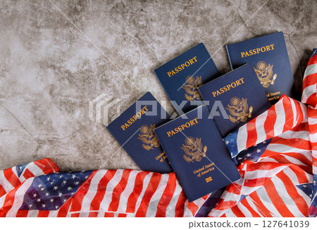 Five United States passports lie on gray surface beside American flag, symbolizing national identity. 127641039