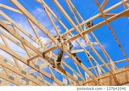 Skilled worker balances on wooden beams while constructing trusses frame of new house Skilled worker balances on wooden beams while constructing trusses frame of new house 127641046