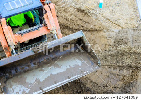 Excavator is digging into earth as construction worker operates t machinery on works day time Excavator is digging into earth as construction worker operates t machinery on works day time 127641069