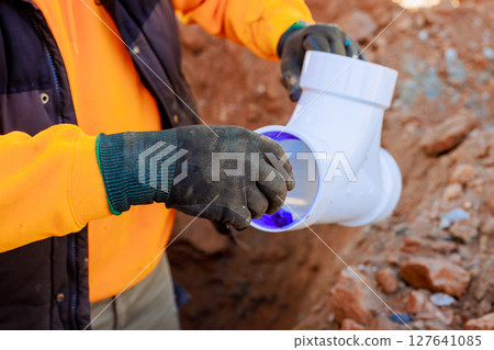 Plumber in gloves connects plumbing fitting sewage pipe while working in trench at construction site during works day 127641085