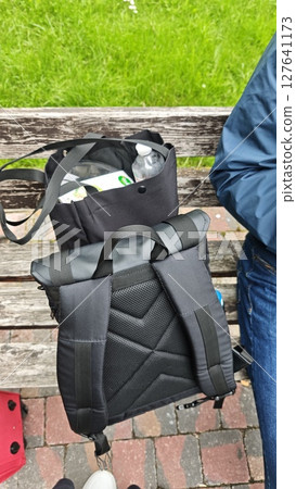 Black backpack and tote bag with travel items placed on a wooden bench in a park. Concept of travel, tourism and outdoor preparation Black backpack and tote bag with travel items placed on a wooden bench in a park. Concept of travel, tourism and outdoor preparation 127641173