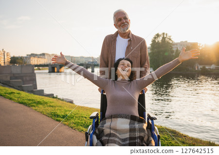 Senior woman in wheelchair walking with caregiver old man on road. Family couple man husband supporting taking care of paralyzed woman wife in chair for people with disability outdoor. Rehabilitation 127642515