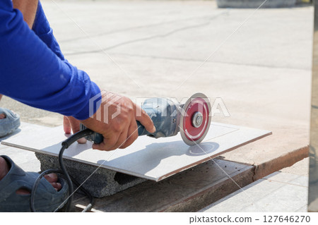 Construction site scene with a worker in blue clothing using an angle grinder to cut a gray tile, highlighting detailed craftsmanship and precision in tile preparation. Construction site scene with a worker in blue clothing using an angle grinder to cut a gray tile, highlighting detailed craftsmanship and precision in tile preparation. 127646270