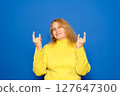 Delighted young ethnic female millennial with long dark hair in warm sweater and sunglasses, smiling brightly and showing rock gesture while listening to favorite song against yellow background 127647300