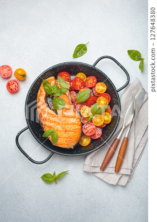 Grilled salmon steak with cherry tomatoes and basil leaves in black pan with cutlery top view on gray background 127648380
