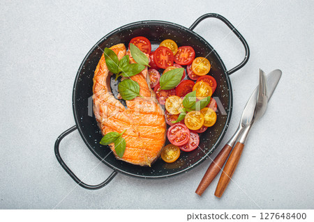Grilled salmon steak with cherry tomatoes and basil leaves in black pan with cutlery top view on gray background Grilled salmon steak with cherry tomatoes and basil leaves in black pan with cutlery top view on gray background 127648400