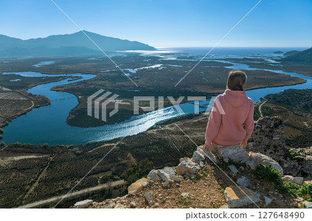 Woman looking from acropolis on top of mount of ancient Greek city Kaunos to Dalyan river, Sulungur lake, Iztuzu beach. Popular place for family travel, boat cruises on summer vacation. Mugla, Turkey Woman looking from acropolis on top of mount of ancient Greek city Kaunos to Dalyan river, Sulungur lake, Iztuzu beach. Popular place for family travel, boat cruises on summer vacation. Mugla, Turkey 127648490