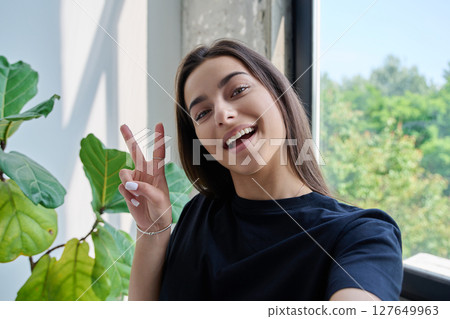 Close-up selfie portrait of teenage female looking at web camera showing victory gesture 127649963