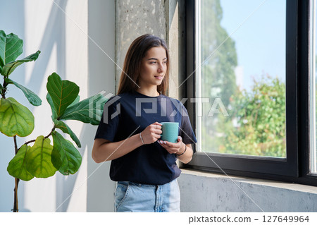 Portrait of beautiful calm relaxed teenage girl with cup in hands at home looking out window 127649964