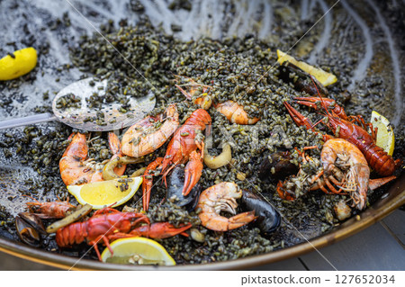 Black seafood paella with shrimp, squid, mussels, lemon wedges served in large pan at food event. Concept of black paella, marine flavors, festive culinary presentation Black seafood paella with shrimp, squid, mussels, lemon wedges served in large pan at food event. Concept of black paella, marine flavors, festive culinary presentation 127652034